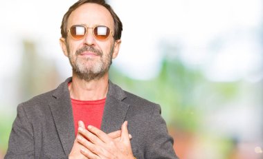 Orta yaş yakışıklı iş adamı ile gülümseyen güneş gözlüğü takıyor kapalı gözler ve yüzünde minnettar jest ile göğüs üzerinde eller. Sağlık kavramı.