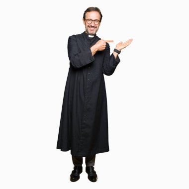 Orta yaş rahip adam hayret ve el ile sunulması ve parmak ile işaret ederken kameraya gülümseyen Katolik bornoz giymiş.