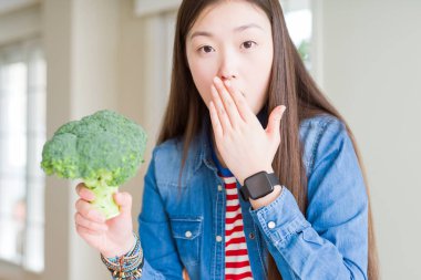 Güzel Asyalı kadın yeşil taze brokoli yemek hata için utanç ile şok el ile ağız kapağı, korku ifadesi, sessizlik, gizli kavram korkmuş
