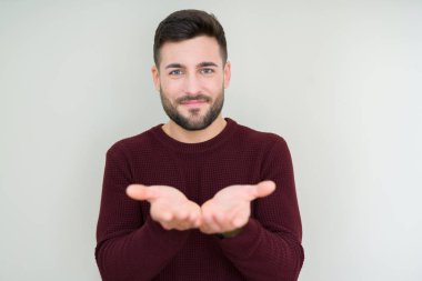 Genç yakışıklı adam izole arka plan üzerinde bir kazak giyen birlikte alma veya jest veren avuç içi ile gülümseyerek. Tutma ve koruma