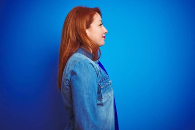Genç güzel kızıl saçlı kadın yan bakarak mavi izole arka plan üzerinde duran denim gömlek giyen, doğal yüz ve kendine güvenen gülümseme ile profil poz dinlenmek.