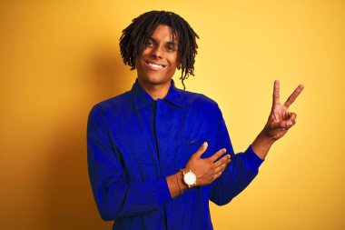 Izole sarı arka plan üzerinde mekanik üniforma giyen dreadlocks ile Afro işçi adam mutlu yüz zafer işareti yapıyor kameraya göz kırparak gülümseyerek. İki numara.
