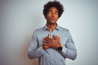 Izole beyaz arka plan üzerinde gri gömlek giyen afro saçlı genç afro-amerikan adam kapalı gözleri ve yüzünde minnettar jest ile göğüs elleri ile gülümseyerek. Sağlık kavramı.