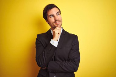 Takım elbise giyen ve kravat takan genç işadamı, elini çenesi üzerinde olan izole sarı arka plan üzerinde durarak soru, dalgın ifade. Gülümseyen ve düşünceli bir yüz. Şüphe kavramı.