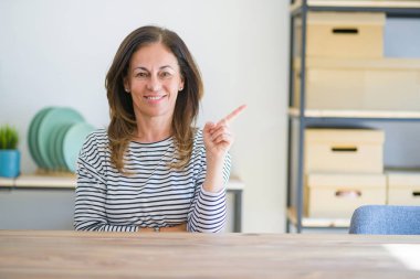 Orta yaş yaşlı kadın yüzünde büyük bir gülümseme ile evde masada oturan, kameraya bakarak yan el parmağı ile işaret.