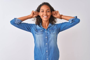 Denim gömlek giyen genç brezilyalı kadın izole beyaz arka plan üzerinde duran gülümseyerek parmakları ile kulakları çekerek, komik jest. Seçme sorunu