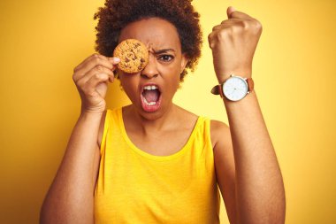 Sarı arka plan üzerinde çikolata cips çerez tutan Afrikalı Amerikalı kadın kızgın ve sinirli öfke, deli ve yükseltilmiş el, öfke kavramı ile bağırarak