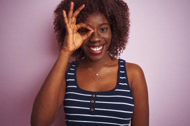 Genç afrikalı afro kadın çizgili t-shirt izole pembe arka plan üzerinde duran el gülümseyerek ok jest yapıyor, mutlu yüz ile parmaklardan bakan göz.