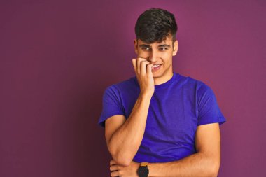 İzole mor arka plan üzerinde duran t-shirt giyen genç hintli adam stresli ve ağız tırnakısırma elleri ile sinirli görünümlü. Anksiyete sorunu.