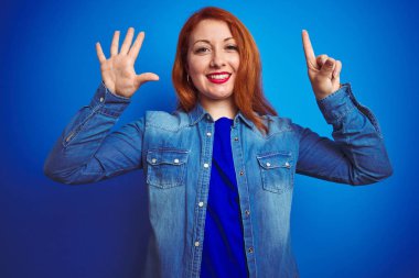 Denim gömlek giyen genç güzel kızıl saçlı kadın mavi izole arka plan üzerinde duran gösteren ve parmakları ile işaret ederken emin ve mutlu gülümseyerek altı numara.