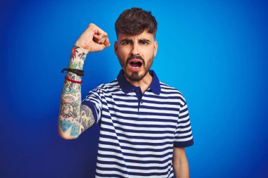 Çizgili polo giyen dövme giyen genç adam izole mavi arka plan üzerinde duran kızgın ve öfkeli öfke ile bağırırken sinirli ve öfkeli yükselterek. Öfke ve agresif kavram.