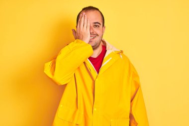 Bir gözünü eliyle kaplayan izole sarı arka plan üzerinde duran yağmurluk giyen genç adam, yüzünde kendine güvenen bir gülümseme ve sürpriz duygular.