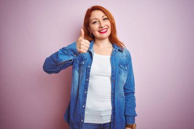 Youg güzel kızıl saçlı kadın izole pembe arka plan üzerinde duran kot gömlek giyen el ile jest kadar mutlu başparmak yapıyor. Başarı gösteren kameraya bakarak ifadeyi onaylama.