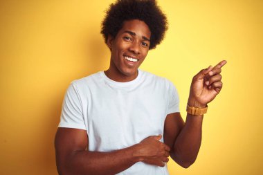 Afro saçlı amerikalı adam beyaz t-shirt giyen yüzünde büyük bir gülümseme ile izole sarı arka plan üzerinde duran, kameraya bakarak yan el ve parmak ile işaret.
