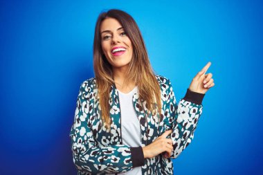 Yüzünde büyük bir gülümseme ile mavi izole arka plan üzerinde duran bir ceket giyen genç güzel kadın, kameraya bakarak yan el ve parmak ile işaret.