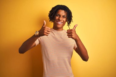 Çizgili t-shirt giyen dreadlocks giyen Afro adam izole sarı arka plan başarı işareti el ile olumlu jest yapıyor, gülümseyen ve mutlu başparmak kadar. Neşeli ifade ve kazanan jest.