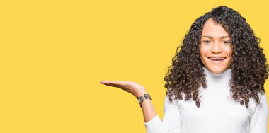 Kıvırcık saçlı, kazak kazak giyen genç güzel kadın neşeli bir şekilde sunarken ve avuç içi kameraya bakarak işaret ederek.