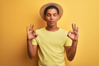 Genç yakışıklı Arap adam izole yelllow arka plan üzerinde t-shirt ve yaz şapka giyen dinlenmek ve gözleri parmakları ile meditasyon jest yapıyor kapalı gülümseyerek. Yoga konsepti.