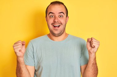 Yeşil rahat t-shirt giyen genç adam izole sarı arka plan üzerinde ayakta sürpriz ve kollarını kaldırdı ve açık gözleri ile başarı için şaşırttı. Kazanan kavramı.