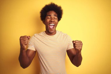 Afro saçlı, çizgili tişört giyen Amerikalı adam izole sarı arka plan üzerinde ayakta kollarını kaldırdı ve gözleri gülümseyerek zafer kutlayan kapalı başarı için heyecanlı. Kazanan kavramı.