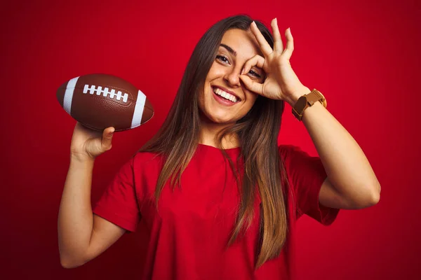 年轻美丽的女运动员拿着足球在孤立的红色背景与快乐的脸微笑做Ok标志手在眼睛通过手指看 — 图库照片