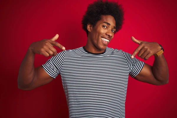 Young american man with afro hair wearing navy striped t-shirt over isolated red background looking confident with smile on face, pointing oneself with fingers proud and happy.