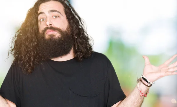 Young man with long hair and beard wearing heavy metal black outfit clueless and confused expression with arms and hands raised. Doubt concept.