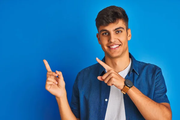 孤立した青い背景の上に立つシャツを着た若いインド人男性が微笑み 両手と指を横に向けてカメラを見ている — ストック写真
