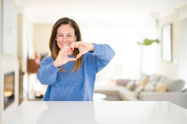 Beautiful middle age woman at home smiling in love showing heart symbol and shape with hands. Romantic concept.