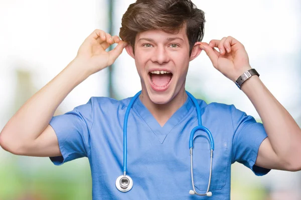 年轻的医生穿着医疗制服在孤立的背景微笑拉耳朵用手指 滑稽的手势 试听问题 — 图库照片
