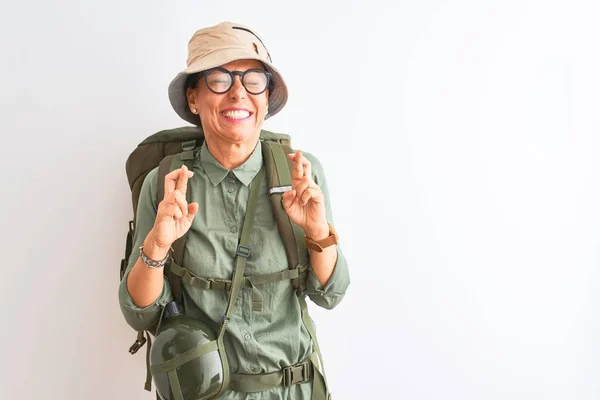 Middle age hiker woman wearing backpack canteen hat glasses over isolated white background gesturing finger crossed smiling with hope and eyes closed. Luck and superstitious concept.