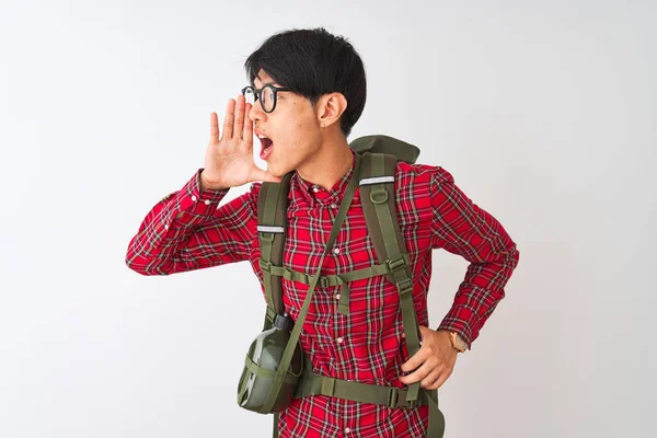 Chinese hiker man wearing backpack canteen glasses over isolated white background shouting and screaming loud to side with hand on mouth. Communication concept.