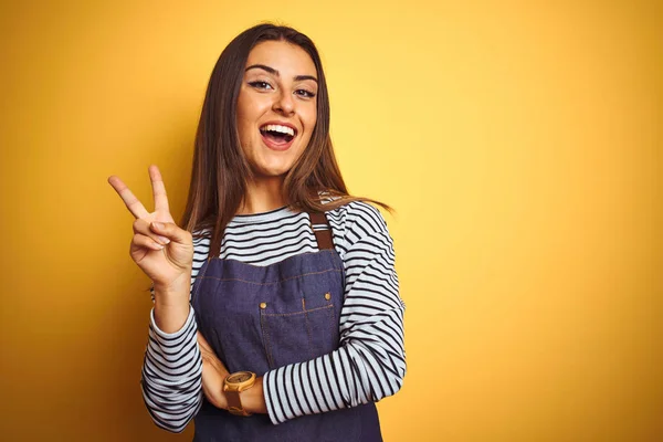 Young beautiful baker woman wearing apron standing over isolated yellow background smiling with happy face winking at the camera doing victory sign. Number two.