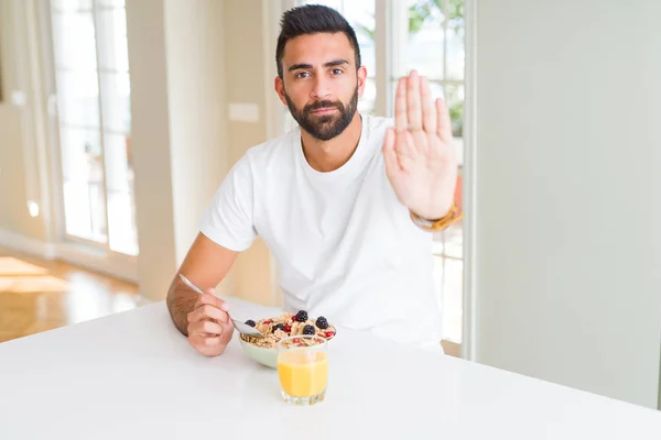 Handsome hispanic man eating healthy breakfast in the morning at home with open hand doing stop sign with serious and confident expression, defense gesture