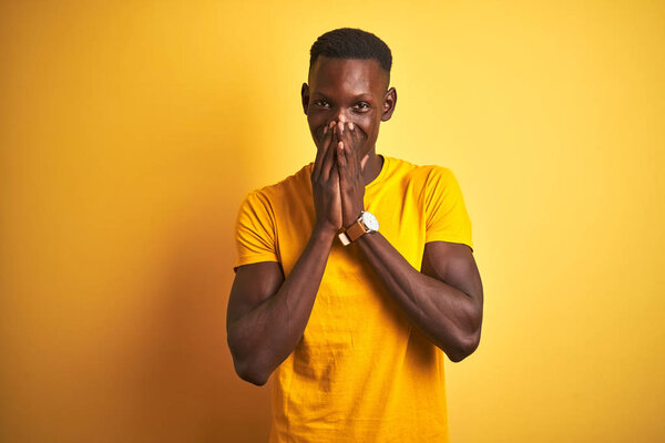 Young african american man wearing casual t-shirt standing over isolated yellow background laughing and embarrassed giggle covering mouth with hands, gossip and scandal concept