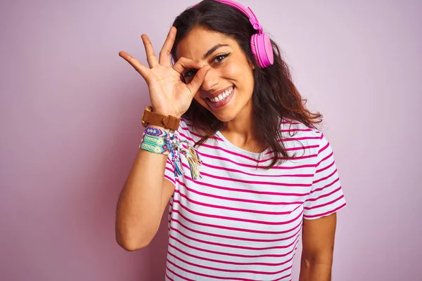年轻的美女听音乐使用耳机在孤立的粉红色背景做确定手势与手微笑 眼睛透过手指与快乐的脸 — 图库照片