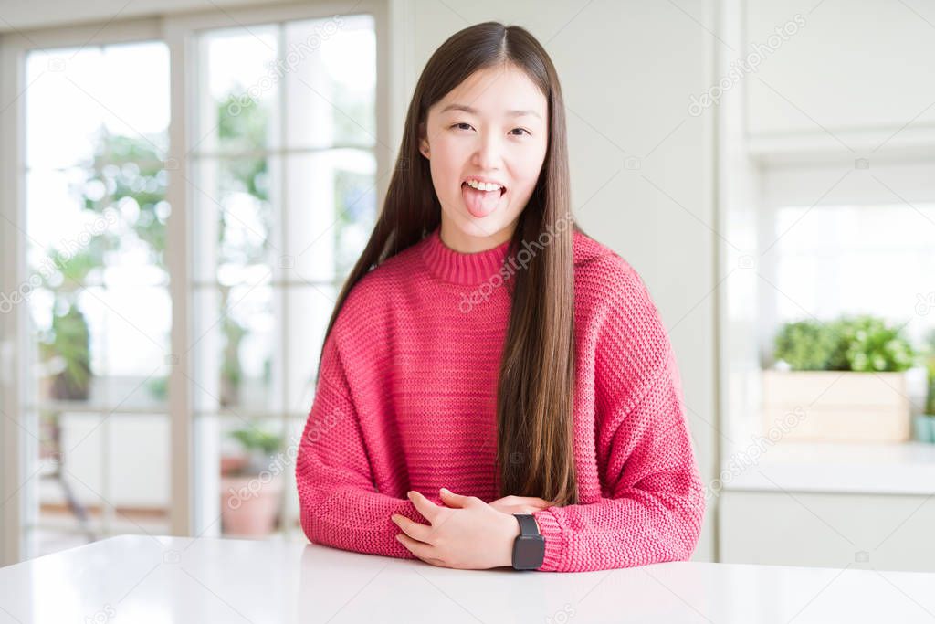 Beautiful Asian woman wearing pink sweater on white table sticking tongue out happy with funny expression. Emotion concept.