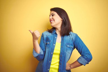 Genç güzel esmer kadın sarı izole arka plan üzerinde rahat kot ceket giyen mutlu yüz görünümlü ve başparmak ile yan işaret gülümseyerek.