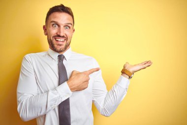 Genç yakışıklı iş adamı sarı izole arka plan üzerinde zarif beyaz gömlek giyen hayran ve el ile sunarken kameraya gülümseyerek ve parmak ile işaret.