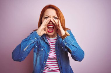 Denim gömlek giyen güzel kızıl saçlı kadın ve izole pembe arka plan üzerinde çizgili t-shirt ağız üzerinde elleri ile öfkeli bağırarak