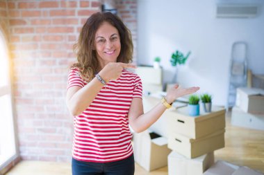 Orta yaş yaşlı kadın yeni bir eve hareketli karton kutular ambalaj şaşırttı ve el ile sunarken kameraya gülümseyerek ve parmak ile işaret.