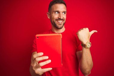 Genç adam kırmızı izole arka plan üzerinde dijital touchpad tablet kullanarak işaret ve mutlu yüz gülümseyerek yan başparmak ile gösteren