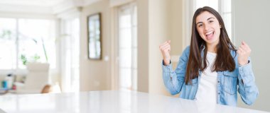 Güzel genç kadın evde beyaz masaya oturan geniş açı resim şaşırmış ve başarı için hayran kaldırdı Kollarını açarak kutluyor ve gözleri açık. Kazanan kavramı.