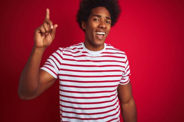 Afro saçlı genç afro-amerikan adam izole kırmızı arka plan üzerinde çizgili t-shirt giyen başarılı bir fikir ile parmak işaret. Çıktı ve mutlu. Bir numara.