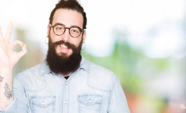 Uzun saç ve sakal el ve parmaklar ok işaretiyle yapıyor olumlu gülümseyen gözlüklü genç hipster adam. Başarılı ifade.