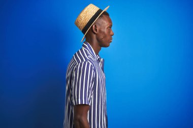 Izole mavi arka plan üzerinde çizgili gömlek ve yazlık şapka giyen Afro-Amerikan adam yan bakarak, kendine güvenen gülümseme ile doğal yüz ile profil poz dinlenmek.
