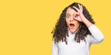 Sürpriz yüz, göz parmaklarının arasından seyir ile ok jest yapıyor boğazlı kazak giyen kıvırcık saçlı genç güzel kadın şok. İmansız ifade.