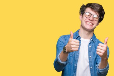 El ile pozitif jest yapıyor onaylama izole arka plan üzerinde gözlük takan genç yakışıklı adam başparmak gülümseyen ve başarı için mutlu. Kamera, kazanan jest arıyorsunuz.