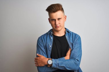 Genç yakışıklı adam izole beyaz arka plan şüpheci ve sinir üzerinde duran denim gömlek giyen, çapraz kolları ile yüzünde onaylamayan ifade. Negatif kişi.