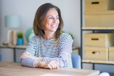 Orta yaş yaşlı kadın evde masada oturan yüzünde gülümseme ile yan bakıyor, doğal ifade. Gülmekten emin.